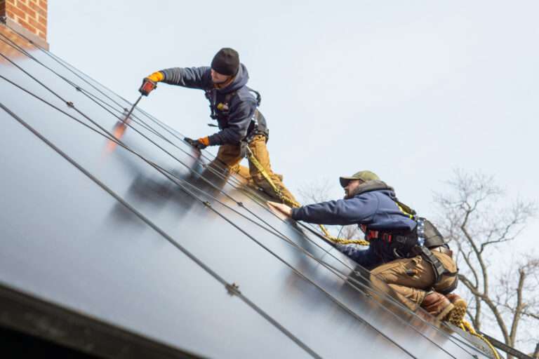 Two electricians in safety gear installing rooftop solar panels on a steep residential roof.